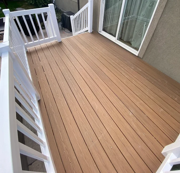 A clean, light brown wooden deck with a sturdy white railing, attached to a house with beige siding and a sliding glass door. The elevated build overlooks a small yard with greenery visible in the background.