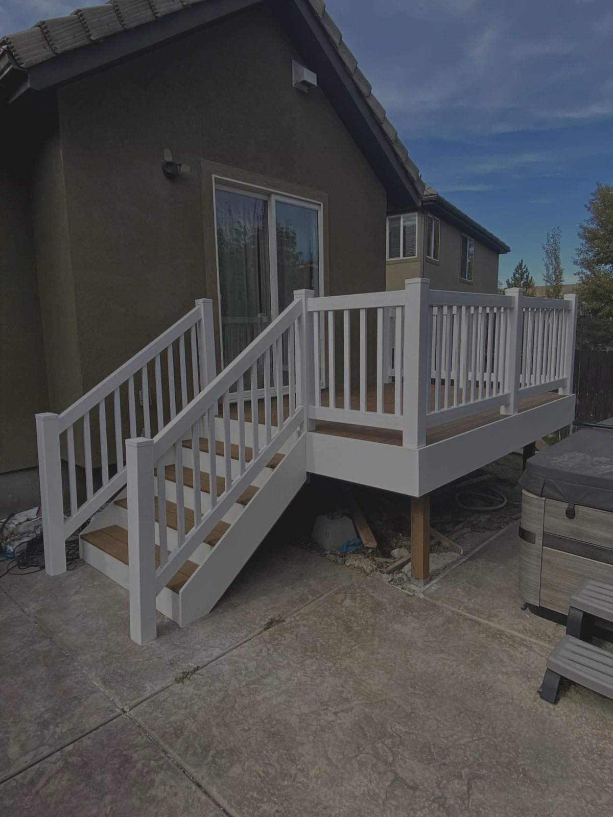 A small backyard deck with white railings and brown steps is attached to a house. The deck has stairs leading down to a concrete patio, with a hot tub and some trees visible nearby.