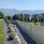 A white vinyl fence separates a landscaped yard with green grass and rocks from neighboring homes, while pine trees and distant mountains rise beyond the fence under a clear blue sky.