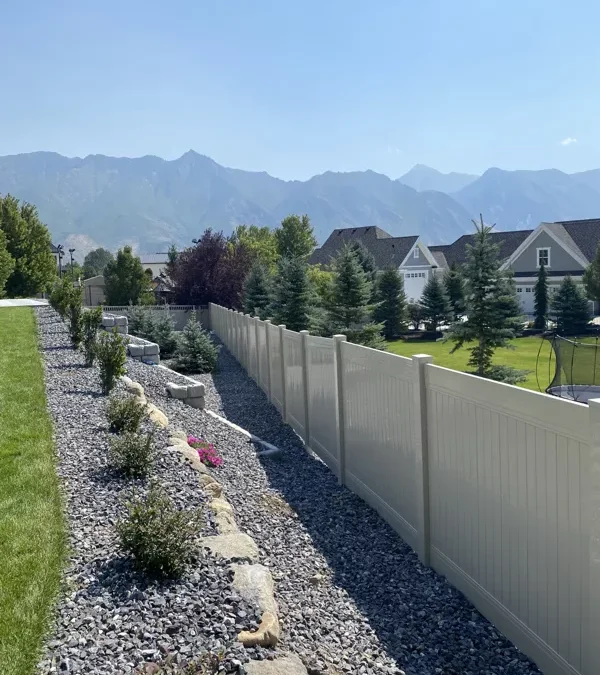 A white vinyl fence separates a landscaped yard with green grass and rocks from neighboring homes, while pine trees and distant mountains rise beyond the fence under a clear blue sky.