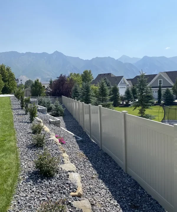 A white vinyl fence separates a landscaped yard with green grass and rocks from neighboring homes, while pine trees and distant mountains rise beyond the fence under a clear blue sky.