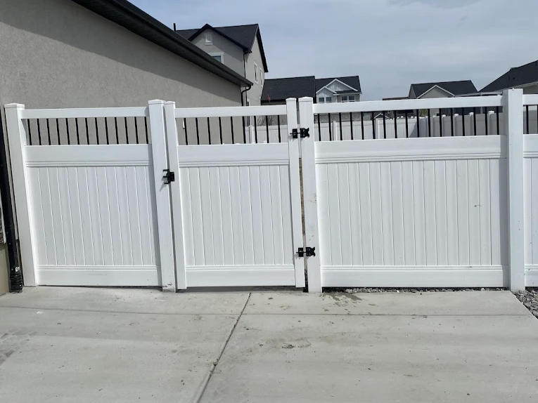 A white vinyl fence with vertical slats and black metal accents stands on a concrete driveway between two houses in a suburban neighborhood.