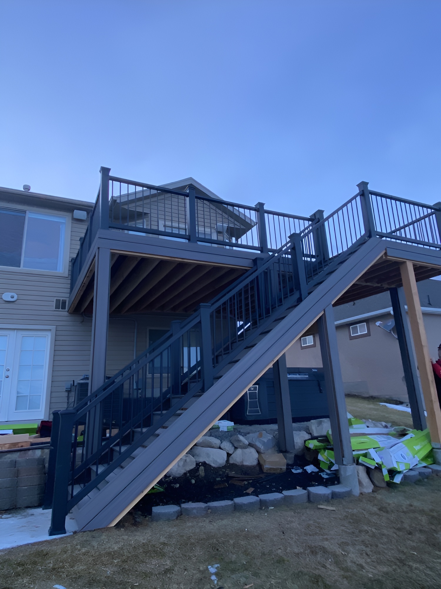 Two-story deck attached to a beige house in Utah, featuring a staircase with dark railings and wooden steps leading to an upper level. The yard below has patchy grass, rocks, and some green tarps or bags.