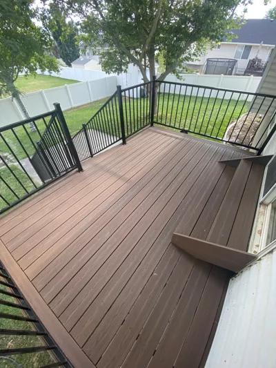 A freshly built deck with black metal railings attached to a house in Utah, overlooking a grassy backyard with trees, white fences, and neighboring houses in the background.