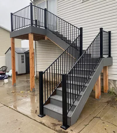 A modern outdoor staircase with sleek black railing and grey steps leads to a raised deck attached to a light-colored Utah home, supported by wooden posts on a wet concrete patio.