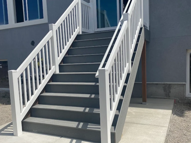 A set of outdoor stairs with dark gray steps and white railings leads up to a doorway on a gray building, situated on a concrete patio.