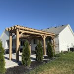 A wooden pergola stands on a concrete patio in a backyard next to a light-colored house. Several small evergreen trees line the patio’s edge, and a fence borders the yard. The sky is clear and blue.